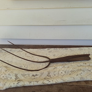 May include: An antique, rusty metal pitchfork with three tines and a wooden handle. The pitchfork is resting on a cream-colored lace tablecloth, with a white wooden background.