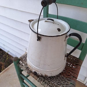 May include: A vintage white enamelware coffee pot with a black handle and rim, resting on a green chair with a woven seat. The pot shows signs of wear, adding to its antique charm. The handle is made of wood.