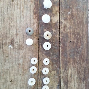 May include: A collection of white ceramic knobs and washers arranged on a weathered wooden surface. The knobs vary in size and shape, including round and cylindrical designs. The washers are flat with central holes.