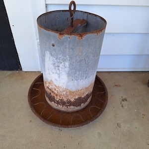 May include: A weathered, cylindrical metal container with a rusty, circular base. The container has a handle and a partially rusted surface, showing shades of gray, white, and brown. The item appears to be an antique or vintage piece.