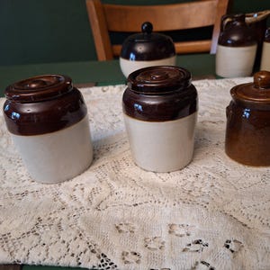 May include: A collection of vintage ceramic jars and containers. The jars are primarily brown and cream-colored, with lids. The arrangement includes jars of varying sizes and shapes, suggesting a set for kitchen or storage use. The jars are displayed on a lace tablecloth.