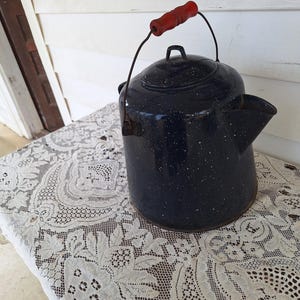 May include: A vintage navy blue enamelware kettle with white speckles. It has a black handle and a red wooden handle. The kettle is sitting on a white lace tablecloth. A classic kitchenware item.
