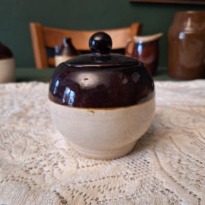 May include: A small, round, antique sugar bowl with a lid. The top half of the bowl and lid are a dark brown, while the bottom half is a cream color. The lid has a small, round knob. The bowl is sitting on a white lace tablecloth.