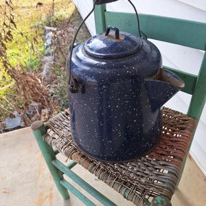 May include: A vintage blue speckled enamelware kettle with a black handle and lid, resting on a green and woven chair. The kettle has a spout and a wire handle. The chair has a distressed, rustic appearance.