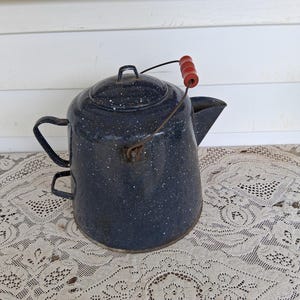 May include: A dark blue enamel coffee pot with a white speckled pattern, featuring a metal bail handle with a red wooden grip, a side handle, a matching lid, and a spout. The pot has a cylindrical body and a metallic rim at the base.