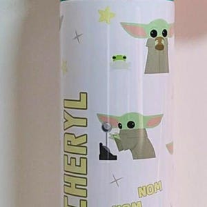 May include: A white cylindrical water bottle with a dark blue and teal flip-top lid. The bottle features a repeating pattern of a green alien character, yellow stars, and the text "NOM NOM" and "Z Z Z". The name "CHERYL" is printed vertically in yellow.