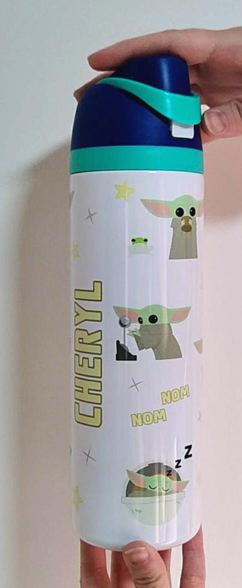 May include: A white cylindrical water bottle with a dark blue and teal flip-top lid. The bottle features a repeating pattern of a green alien character, yellow stars, and the text "NOM NOM" and "Z Z Z". The name "CHERYL" is printed vertically in yellow.
