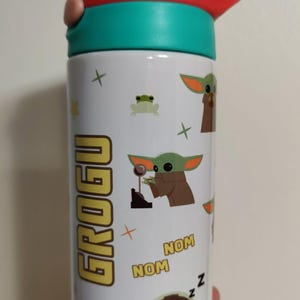 K&ouml;nnte beinhalten: Eine wei&szlig;e Trinkflasche mit einem t&uuml;rkisfarbenen und roten Deckel. Die Flasche ist mit Grogu-Charaktergrafiken verziert, darunter der Schriftzug GROGU, das zweimal wiederholte Wort NOM sowie kleine gr&uuml;ne Fr&ouml;sche, Sterne und Pluszeichen.