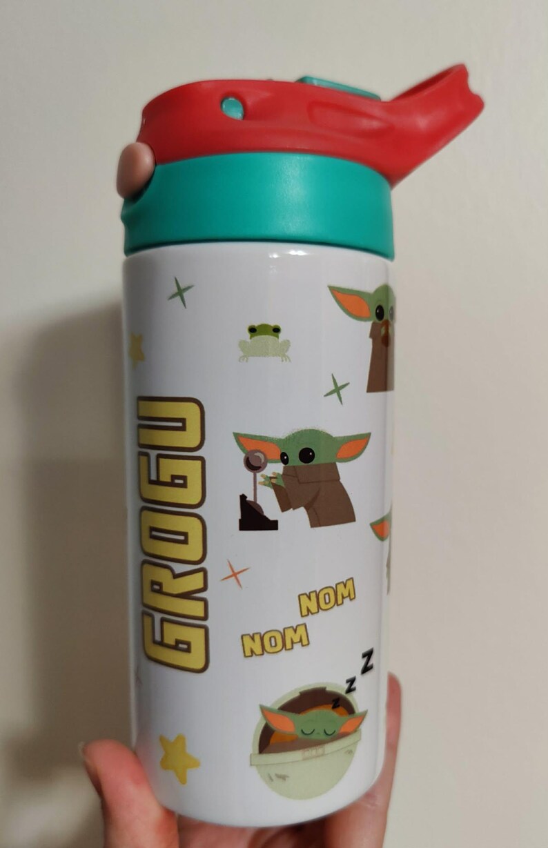 K&ouml;nnte beinhalten: Eine wei&szlig;e Trinkflasche mit einem t&uuml;rkisfarbenen und roten Deckel. Die Flasche ist mit Grogu-Charaktergrafiken verziert, darunter der Schriftzug GROGU, das zweimal wiederholte Wort NOM sowie kleine gr&uuml;ne Fr&ouml;sche, Sterne und Pluszeichen.