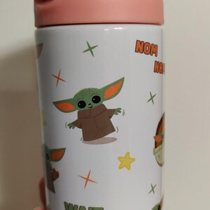 K&ouml;nnte beinhalten: Eine wei&szlig;e zylindrische Trinkflasche mit einem wiederkehrenden Muster aus einer gr&uuml;nen Alien-Figur mit gro&szlig;en Ohren in einer braunen Robe. Die Flasche zeigt gelbe Sterne, orangefarbene Kreuze sowie den Text "NOM NOM" und "WAIT FOR ME!" mit einem violetten Deckel.