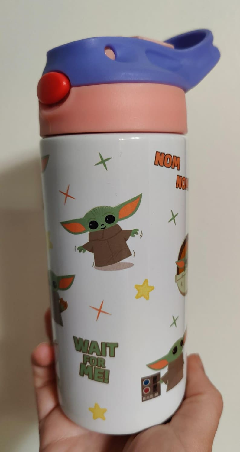 K&ouml;nnte beinhalten: Eine wei&szlig;e zylindrische Trinkflasche mit einem wiederkehrenden Muster aus einer gr&uuml;nen Alien-Figur mit gro&szlig;en Ohren in einer braunen Robe. Die Flasche zeigt gelbe Sterne, orangefarbene Kreuze sowie den Text "NOM NOM" und "WAIT FOR ME!" mit einem violetten Deckel.