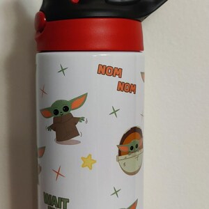 K&ouml;nnte beinhalten: Eine wei&szlig;e Trinkflasche mit einem roten und schwarzen Deckel inklusive Trinkhalm. Die Flasche ist mit Cartoon-Illustrationen einer gr&uuml;nen Kreatur in brauner Robe, gelben Sternen und kleinen Kreuzen verziert. Der Text lautet NOM NOM und WAIT FOR ME.