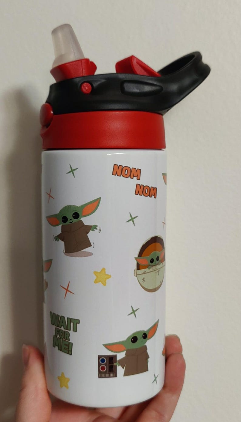 K&ouml;nnte beinhalten: Eine wei&szlig;e Trinkflasche mit einem roten und schwarzen Deckel inklusive Trinkhalm. Die Flasche ist mit Cartoon-Illustrationen einer gr&uuml;nen Kreatur in brauner Robe, gelben Sternen und kleinen Kreuzen verziert. Der Text lautet NOM NOM und WAIT FOR ME.