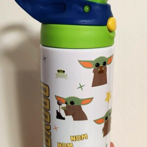K&ouml;nnte beinhalten: Eine wei&szlig;e Trinkflasche mit blau-gr&uuml;nem Deckel und transparentem Trinkhalm. Die Flasche ist mit einem Muster aus der Figur Grogu, einem kleinen gr&uuml;nen Frosch, gelben Sternen sowie dem gelb-schwarzen Text "NOM NOM" und "Z Z Z" bedruckt.