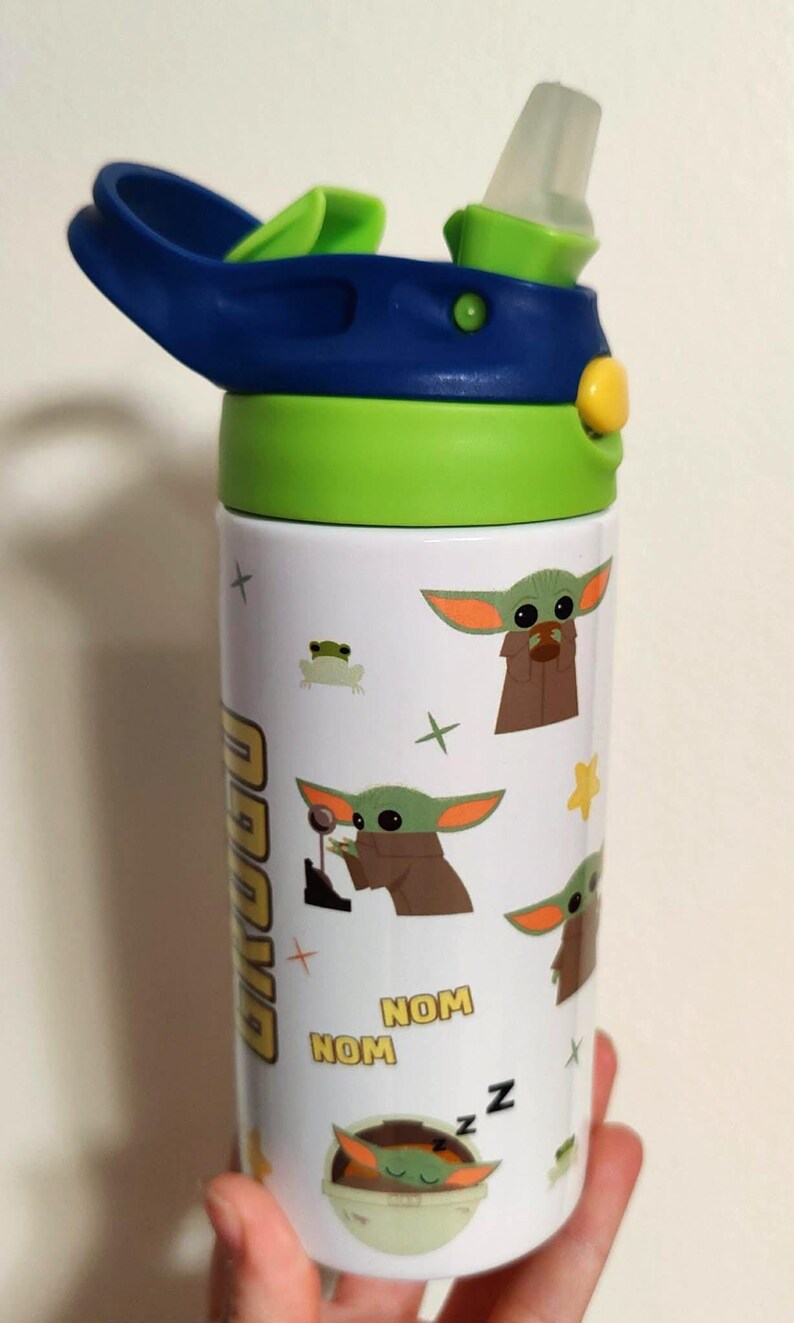 K&ouml;nnte beinhalten: Eine wei&szlig;e Trinkflasche mit blau-gr&uuml;nem Deckel und transparentem Trinkhalm. Die Flasche ist mit einem Muster aus der Figur Grogu, einem kleinen gr&uuml;nen Frosch, gelben Sternen sowie dem gelb-schwarzen Text "NOM NOM" und "Z Z Z" bedruckt.