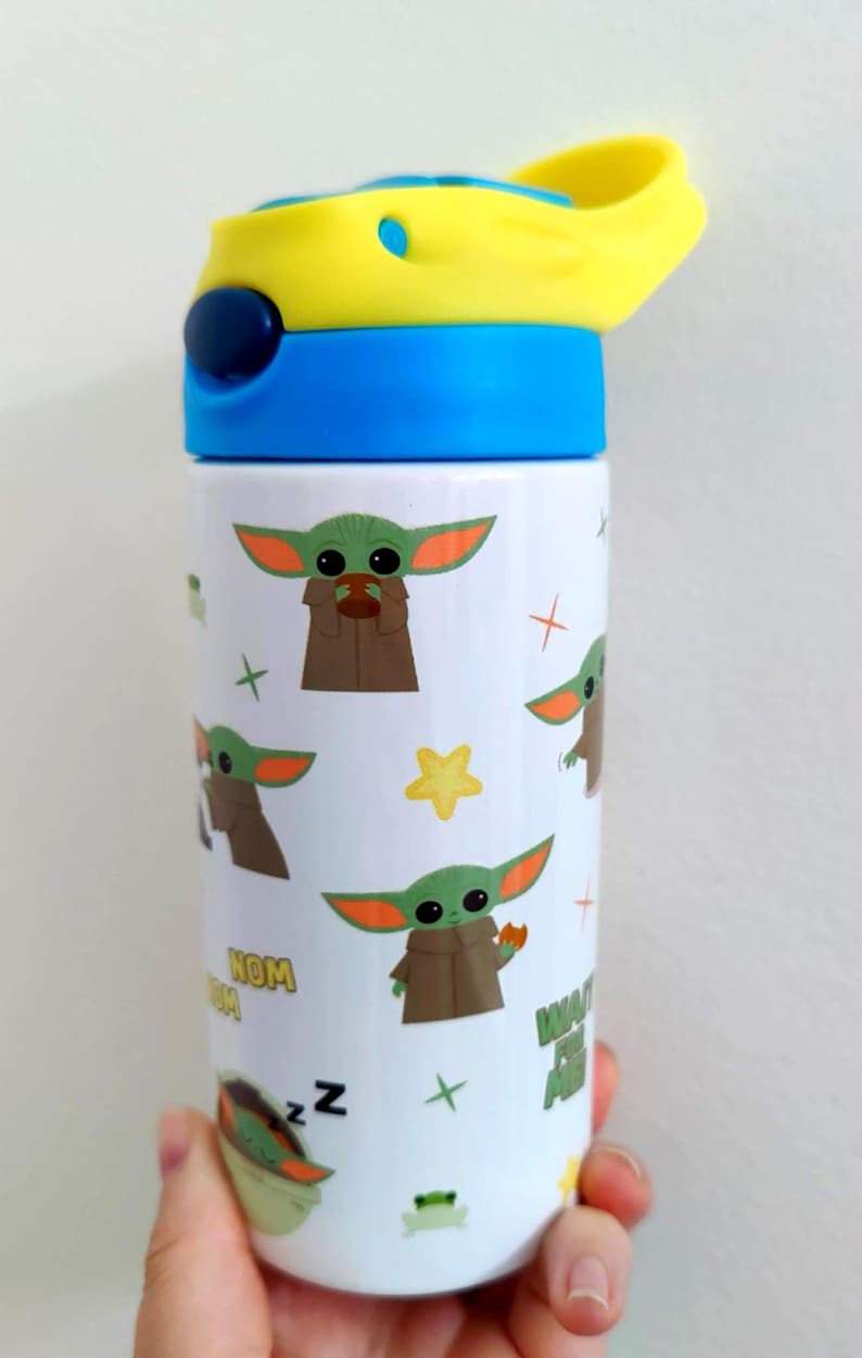 K&ouml;nnte beinhalten: Eine wei&szlig;e zylindrische Trinkflasche mit einem wiederkehrenden Muster aus einer gr&uuml;nen Alien-Figur mit gro&szlig;en Ohren, kleinen Fr&ouml;schen, gelben Sternen und dem Text "NOM NOM". Die Flasche hat einen blauen Deckel mit gelbem Klappverschluss.