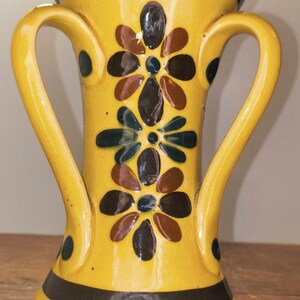 May include: Yellow ceramic vase with two handles, decorated with floral patterns in brown, black, and blue. The vase has a dark band at the base and rim, and is suitable for home decor.