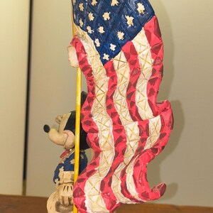 Puede incluir: Una figura patri&oacute;tica con Mickey Mouse sosteniendo una gran bandera estadounidense. La bandera es roja, blanca y azul, con estrellas y rayas. Mickey viste un atuendo patri&oacute;tico. La base es azul con un patr&oacute;n de estrellas.
