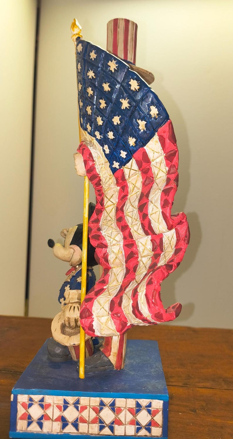 Puede incluir: Una figura patri&oacute;tica con Mickey Mouse sosteniendo una gran bandera estadounidense. La bandera es roja, blanca y azul, con estrellas y rayas. Mickey viste un atuendo patri&oacute;tico. La base es azul con un patr&oacute;n de estrellas.