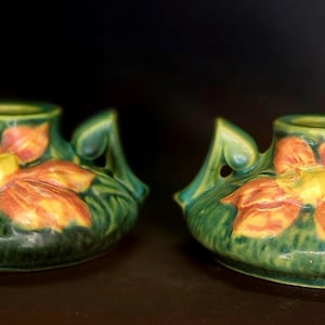 May include: Two matching green ceramic vases with orange and yellow floral designs. Each vase has a rounded base, a central opening, and stylized leaf-shaped handles. The vases are displayed on a dark surface.