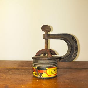May include: A vintage metal clamp with a rusty screw mechanism is holding a small, cylindrical jar. The jar has a yellow and black label with text. The clamp is marked "MADE IN USA". The items are on a wooden surface.