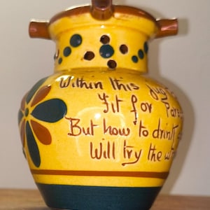 May include: A yellow ceramic jug with a spout and two handles. The jug has a dark blue base and a brown stripe. It features a floral design and text that reads "Within this fit for..." in brown script. Dark blue dots are also on the jug.