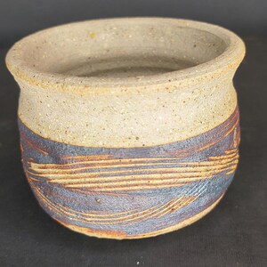 May include: A small, round ceramic pot with a wide opening and a thick, flared rim. The upper portion is a speckled, light beige color, while the lower section features a dark blue glaze with horizontal, textured streaks of exposed brown clay.