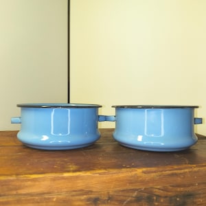 May include: Two light blue enamel pots with black rims and handles. The pots are sitting on a wooden surface. The pots have a classic design and are likely used for cooking.