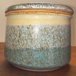 May include: A handcrafted ceramic jar with a lid. The jar features a textured surface with bands of color: a light beige band, a speckled turquoise band, and a darker turquoise base. The lid has a light brown rim.