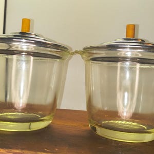 May include: Two vintage glass containers with silver-colored lids and small wooden handles. The containers have a yellow tint and are sitting on a wooden surface. The lids have a handle on top and small handles on the sides.