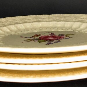 May include: A stack of four cream-colored ceramic plates. Each plate has a textured rim and a central floral design with pink and red roses. The plates are stacked neatly, showcasing their detailed design.