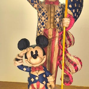 Puede incluir: Una figura patri&oacute;tica con el T&iacute;o Sam y Mickey Mouse con atuendos rojos, blancos y azules. El T&iacute;o Sam saluda mientras sostiene una bandera. Mickey Mouse tambi&eacute;n saluda. La base dice "From Sea To Shining Sea."