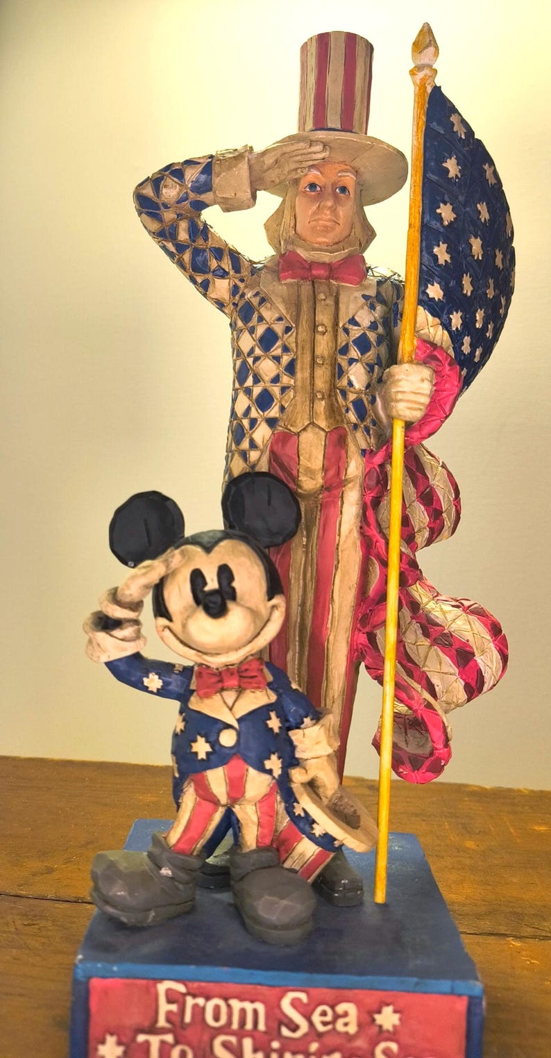 Puede incluir: Una figura patri&oacute;tica con el T&iacute;o Sam y Mickey Mouse con atuendos rojos, blancos y azules. El T&iacute;o Sam saluda mientras sostiene una bandera. Mickey Mouse tambi&eacute;n saluda. La base dice "From Sea To Shining Sea."
