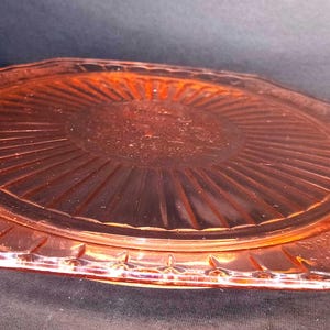 May include: A translucent pink glass serving platter with an octagonal shape and integrated side handles. The center features a sunburst pattern radiating from a circular middle, surrounded by a decorative embossed floral border along the rim.
