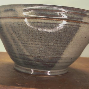 May include: A handmade ceramic bowl with a wide, angled design. The bowl features a mottled gray and brown glaze with a darker band around the rim. The interior is a lighter gray, and the bowl sits on a wooden surface.
