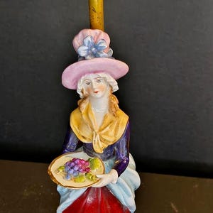 May include: A vintage porcelain figurine lamp base featuring a person wearing a pink hat with a blue flower, a yellow shawl, a dark blue bodice, and a red and white skirt. The figure holds a small plate of grapes atop a textured gold base.