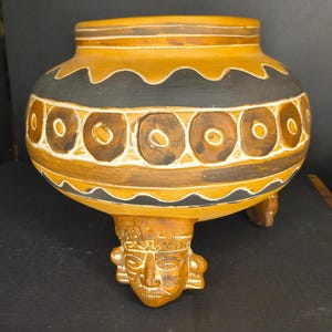 May include: A round, tan ceramic pot featuring three legs sculpted into human faces. The vessel is decorated with horizontal bands of black and white, including a central row of dark brown circles outlined in white and wavy black lines.