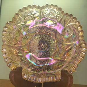 May include: An iridescent, antique glass bowl with a scalloped rim. The bowl features a detailed, geometric pattern with star and floral designs. The glass has a rainbow sheen, reflecting colors of pink, yellow, and blue. The bowl is displayed on a wooden stand.