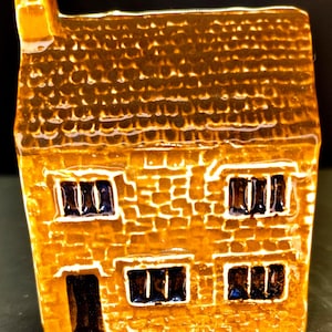 May include: A miniature ceramic house with a textured, golden-brown exterior. The house features a chimney, a dark door, and windows with dark blue interiors. The roof has a detailed, textured pattern.