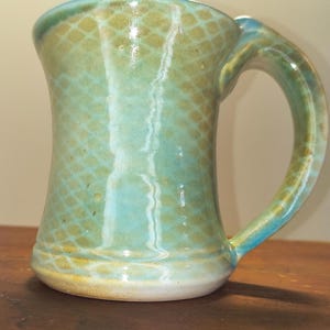 May include: A handmade ceramic mug with a unique design. The mug features a light blue and green glaze with a textured pattern. The handle is a matching color. The mug is approximately 5 inches tall.