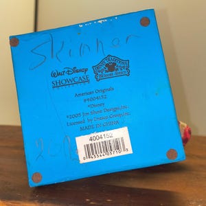 Puede incluir: Base azul de una figura de la colecci&oacute;n Disney Showcase con el texto "Skinner" escrito con rotulador azul. La base incluye el texto "Disney Traditions" y "American Originals #4004152". La figura es de 2005 y est&aacute; hecha en China.