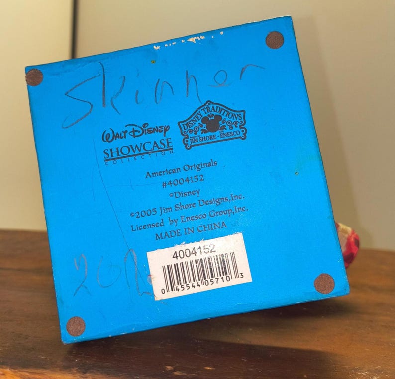 Puede incluir: Base azul de una figura de la colecci&oacute;n Disney Showcase con el texto "Skinner" escrito con rotulador azul. La base incluye el texto "Disney Traditions" y "American Originals #4004152". La figura es de 2005 y est&aacute; hecha en China.