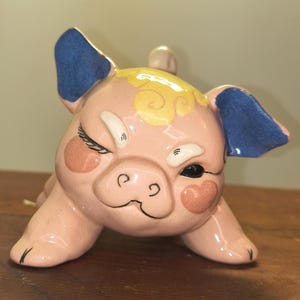May include: A ceramic pig figurine in a playful pose, winking with one eye. The pig is primarily pink with blue ears, yellow accents, and heart-shaped blush. The figurine is on a wooden surface.