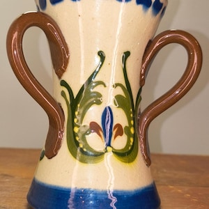 May include: A ceramic vase with two brown handles and a cream-colored body. The vase features a blue base and rim, with green and yellow floral designs. Blue dots decorate the top rim.