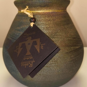 May include: A rounded ceramic jar with a textured, dark metallic finish featuring green and bronze tones. A black square tag hangs from the neck by a string, displaying a gold bird graphic and the text Spirit Art Jar Raku.