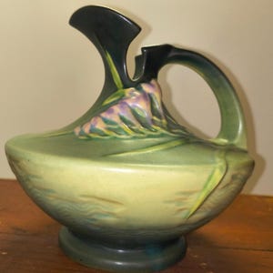 May include: A green ceramic pitcher with a wide base and a curved handle. The pitcher features a floral design in shades of purple and yellow. The spout is black. The pitcher is likely for decorative use.