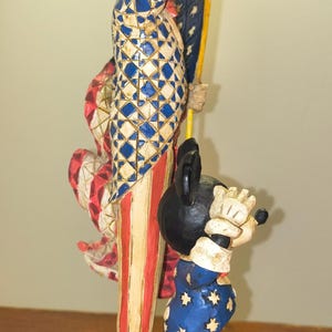 Puede incluir: Una figura de madera tallada que representa al T&iacute;o Sam y Mickey Mouse. El T&iacute;o Sam viste de rojo, blanco y azul, sosteniendo una bandera. Mickey Mouse lleva un traje azul con estrellas blancas. La base es azul con un patr&oacute;n rojo, blanco y azul.