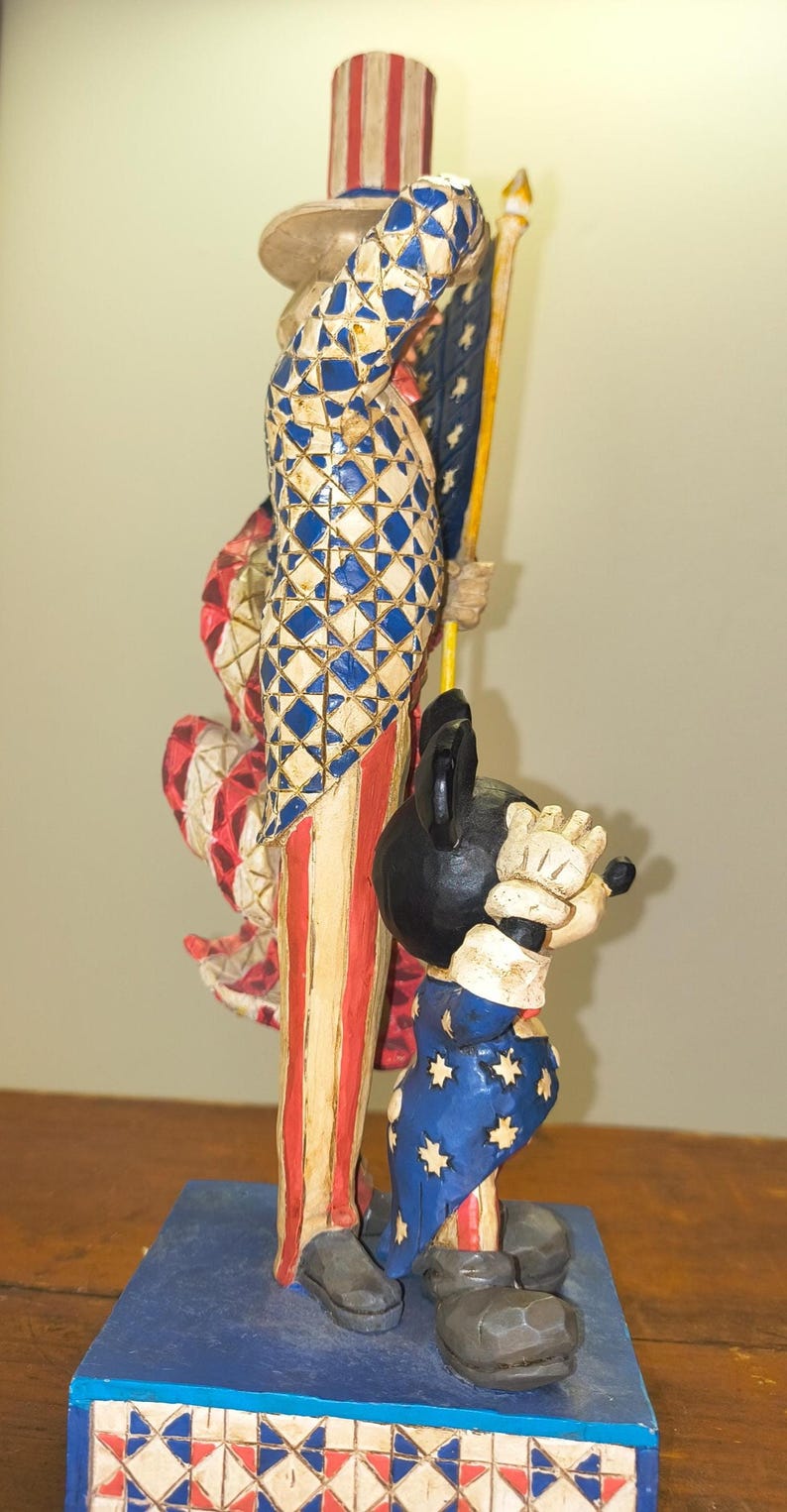 Puede incluir: Una figura de madera tallada que representa al T&iacute;o Sam y Mickey Mouse. El T&iacute;o Sam viste de rojo, blanco y azul, sosteniendo una bandera. Mickey Mouse lleva un traje azul con estrellas blancas. La base es azul con un patr&oacute;n rojo, blanco y azul.