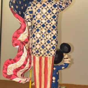 Puede incluir: Una figura patri&oacute;tica que presenta una figura con un atuendo y sombrero de copa con estrellas, sosteniendo una bandera. La figura es roja, blanca y azul. Tambi&eacute;n hay una figura m&aacute;s peque&ntilde;a con un atuendo azul con estrellas blancas. La base es azul con detalles en rojo y blanco.