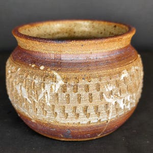 May include: A small, round, hand-thrown ceramic pot with a wide opening and a thick, flared rim. The exterior features a textured, stamped pattern around the center, finished in earthy shades of brown and tan with subtle white glaze accents.